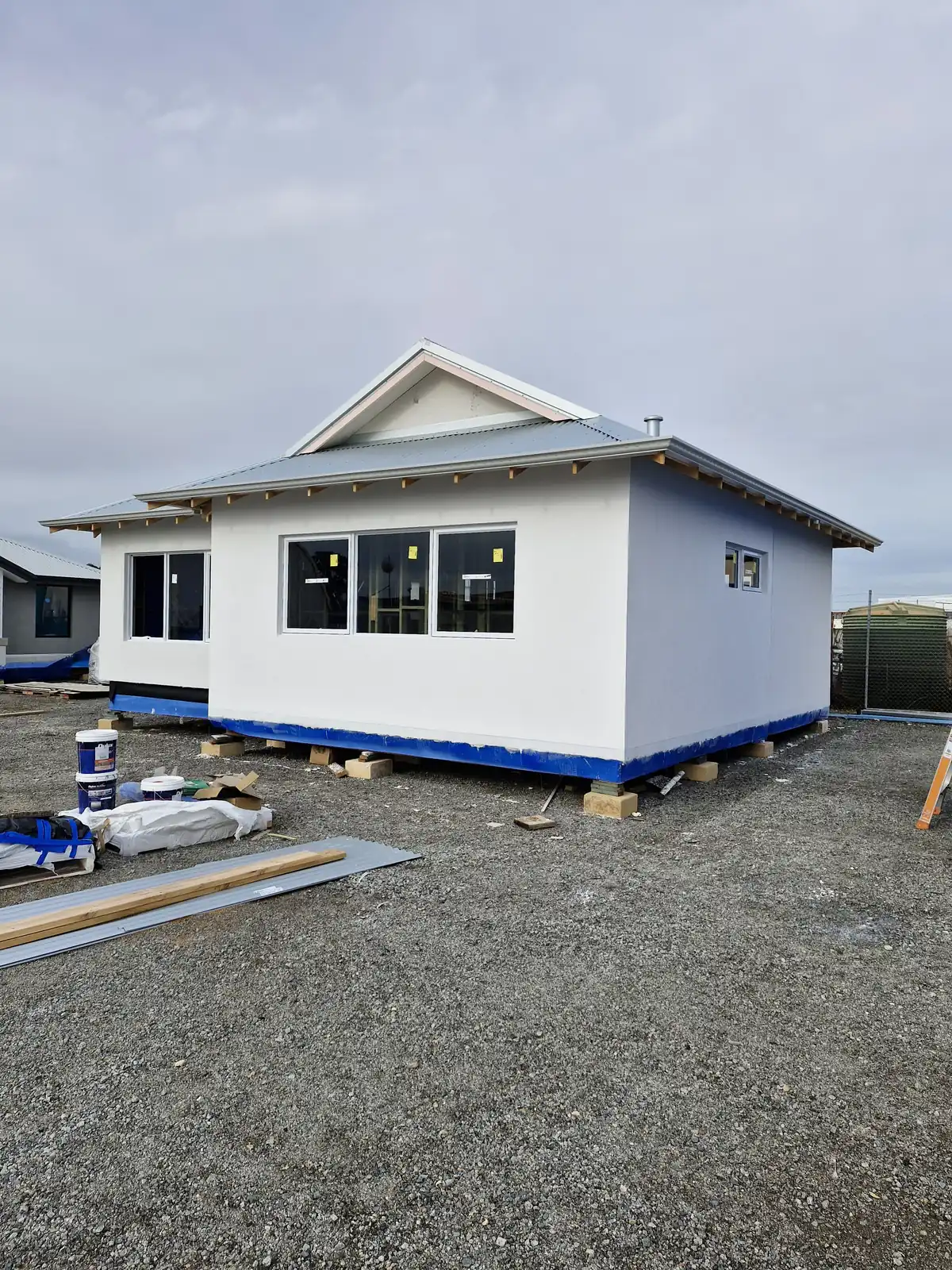 White rendered single-storey building with a grey roof and blue wrap remaining along the base during construction.