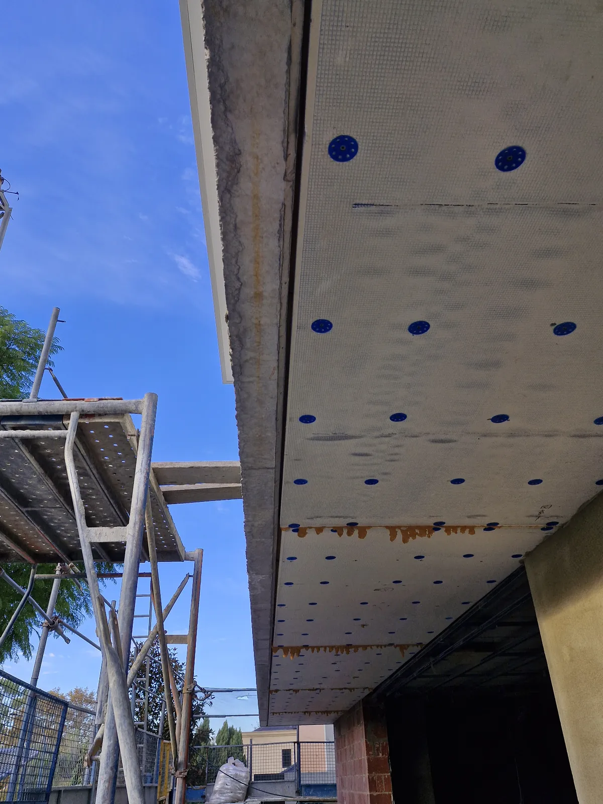 Underside of pale facade panels showing mesh, seams and blue fixing heads beside scaffolding.